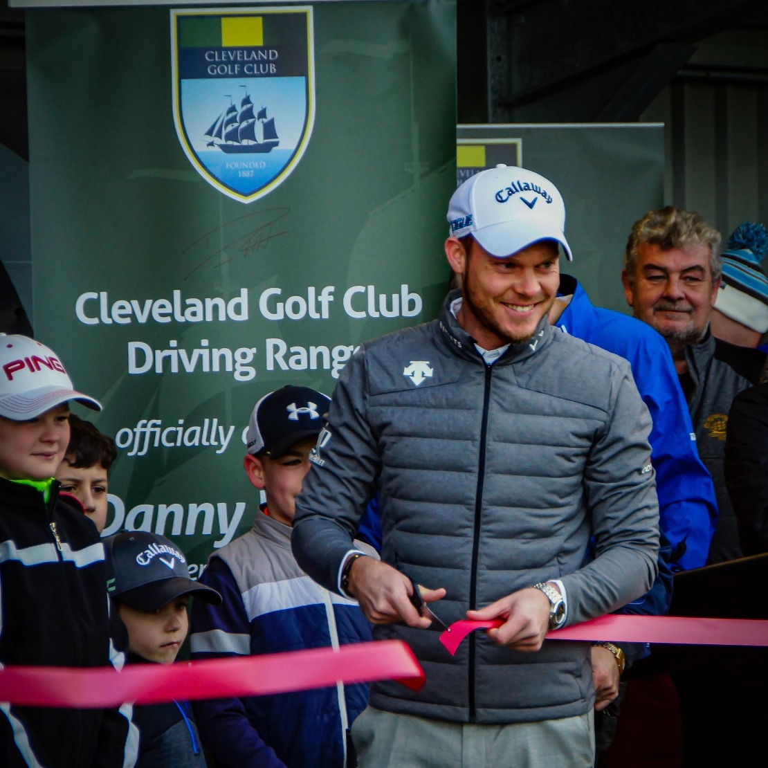 Danny Willett smiling as he cuts the ribbon at the opening of the Cleveland Golf Club driving range, in front of the club's banner